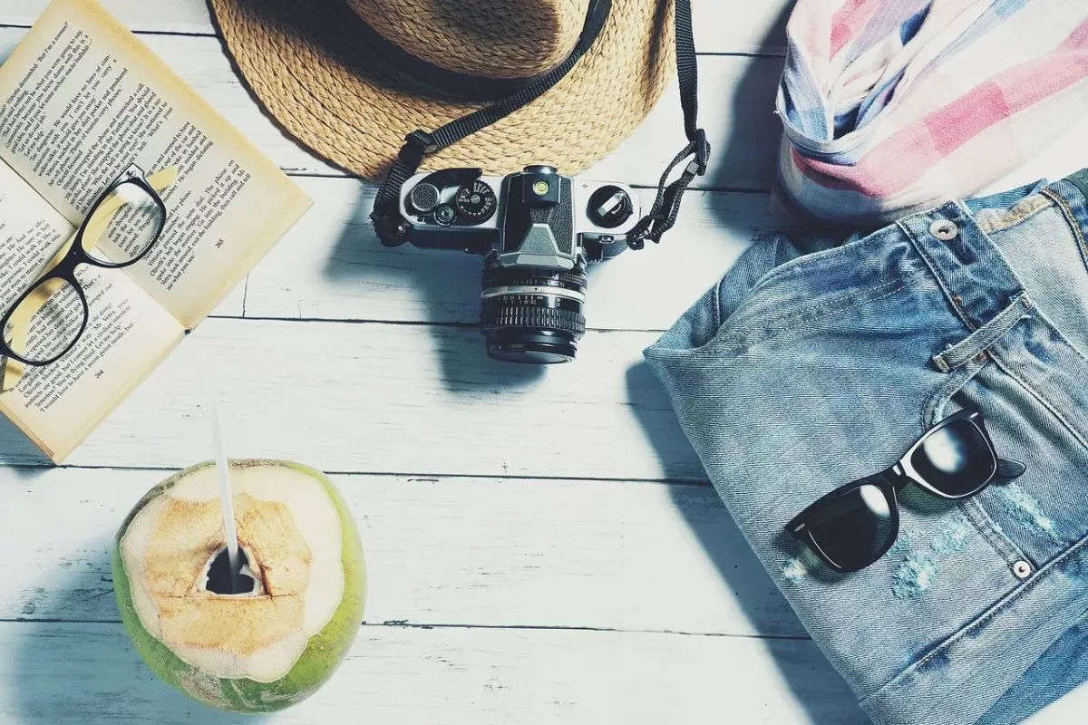 Cette image regroupe un short en jean, un appareil photo, des lunettes de soleils, un livre, une boisson dans une noix de coco, un chapeau rappelant les vacances